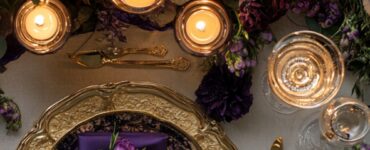 A flat-lay of a wedding setup featuring a purple and gold color scheme, with purple flowers, gold-accented candles, and luxurious gold tableware, capturing a regal and elegant ambiance.