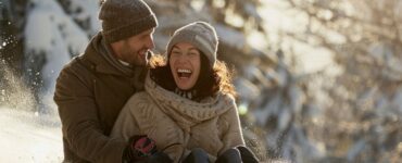 A couple sledding down a snowy hill, laughing and having fun, with snow-covered trees in the background.