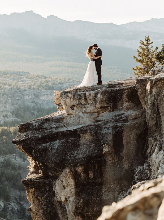 A couple posing for wedding photos on a rocky cliff with a panoramic mountain view, capturing the natural beauty of the surrounding landscape.