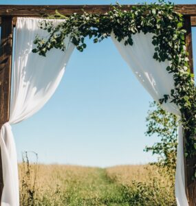 A rustic wedding ceremony setup with a wooden arch draped in white fabric and greenery, surrounded by an open field under a clear blue sky.