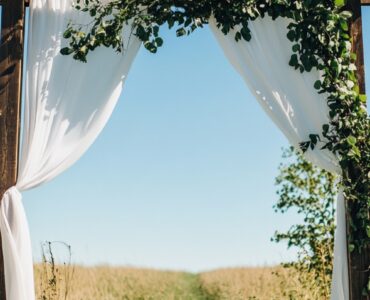 A rustic wedding ceremony setup with a wooden arch draped in white fabric and greenery, surrounded by an open field under a clear blue sky.