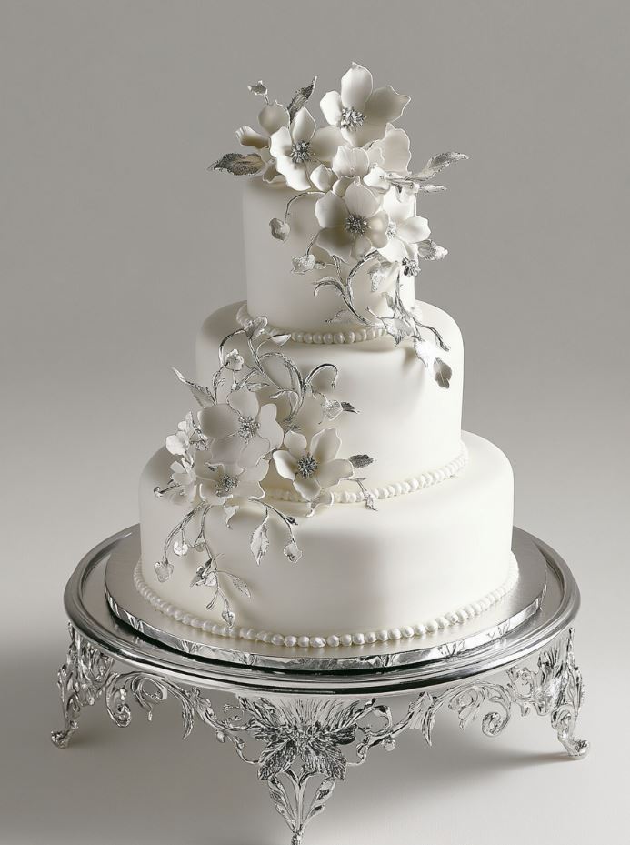 A stunning white wedding cake adorned with silver edible accents, delicate sugar flowers, and displayed on an elegant silver stand.
