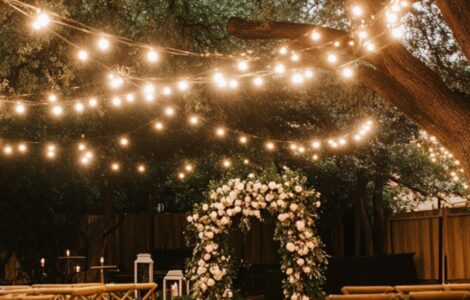 A beautifully decorated backyard wedding ceremony with an intimate setup, including string lights, floral arrangements, and wooden chairs under a large tree.