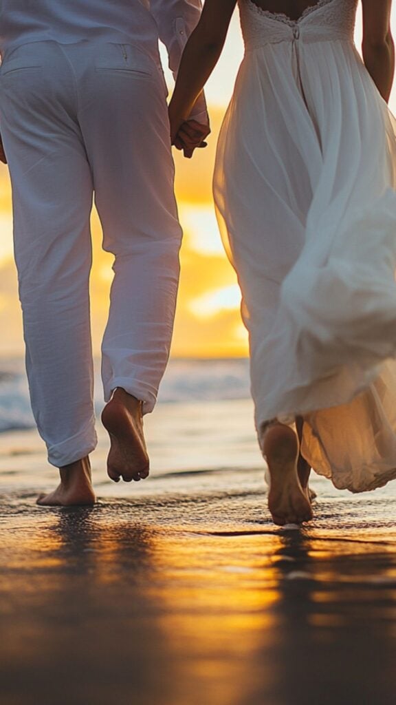 A bride and groom walking barefoot on a sandy beach, with waves gently lapping at their feet and a sunset on the horizon.