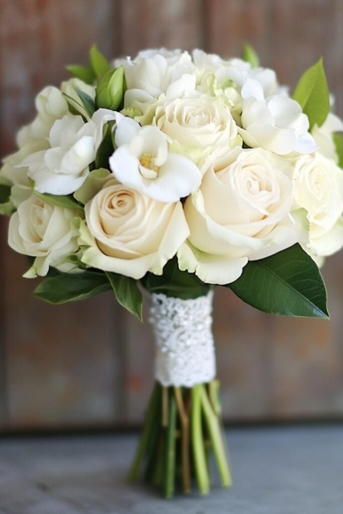A small hand-tied bridal bouquet featuring white roses, gardenias, and subtle greenery wrapped in lace ribbon.