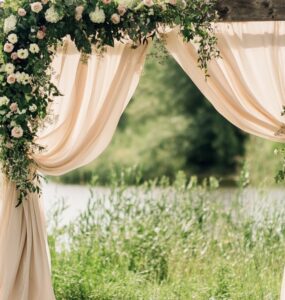 Rustic wedding arch with draped fabrics, florals, and greenery set outdoors, creating an elegant and timeless ceremony backdrop.
