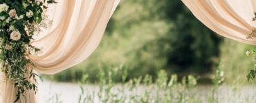 Rustic wedding arch with draped fabrics, florals, and greenery set outdoors, creating an elegant and timeless ceremony backdrop.