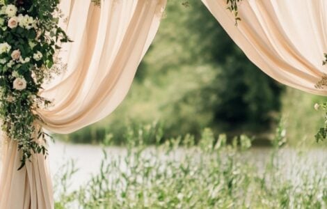 Rustic wedding arch with draped fabrics, florals, and greenery set outdoors, creating an elegant and timeless ceremony backdrop.