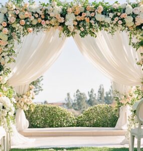 An elegant outdoor spring wedding ceremony setup featuring pastel florals, greenery, and soft draped linens under clear skies.