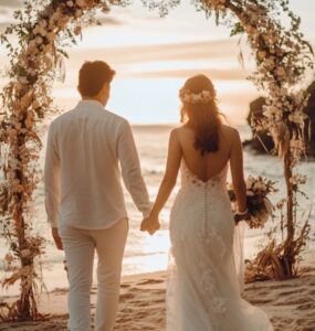 A newlywed couple holding hands, walking barefoot on a beach at sunset, with soft floral décor and a boho-inspired arch in the background.