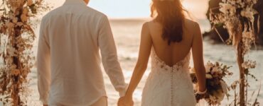 A newlywed couple holding hands, walking barefoot on a beach at sunset, with soft floral décor and a boho-inspired arch in the background.