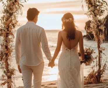 A newlywed couple holding hands, walking barefoot on a beach at sunset, with soft floral décor and a boho-inspired arch in the background.