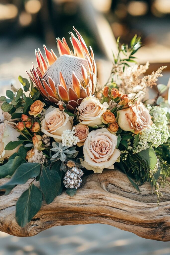 A driftwood centerpiece adorned with floral garlands of roses, proteas, and greenery, accented with feathers and crystals for a beachy boho vibe.