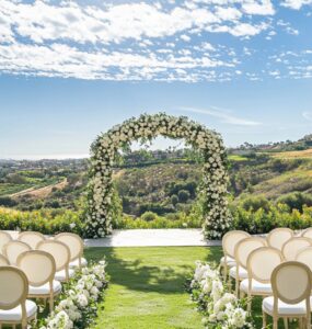A romantic outdoor wedding ceremony setup with a floral arch and white chairs under a sunny sky, surrounded by lush greenery, setting the perfect ambiance for exchanging vows.