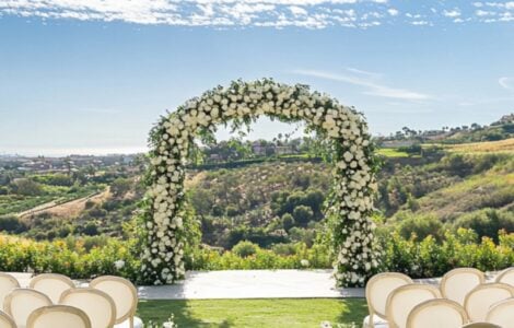 A romantic outdoor wedding ceremony setup with a floral arch and white chairs under a sunny sky, surrounded by lush greenery, setting the perfect ambiance for exchanging vows.