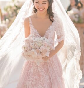 Bride in a soft pink gown walking down the aisle, surrounded by guests, with a radiant smile and a long flowing veil trailing behind.