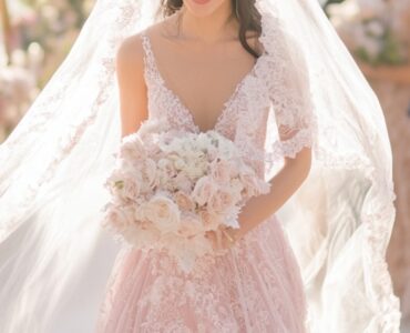 Bride in a soft pink gown walking down the aisle, surrounded by guests, with a radiant smile and a long flowing veil trailing behind.