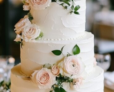 A three-tier wedding cake with soft white buttercream, adorned with blush roses and delicate greenery on a romantic wedding table.