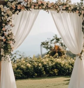 A serene outdoor wedding setup with a simple wooden arch draped in white fabric and adorned with flowers, surrounded by natural greenery under a clear sky.