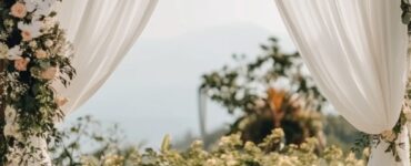 A serene outdoor wedding setup with a simple wooden arch draped in white fabric and adorned with flowers, surrounded by natural greenery under a clear sky.