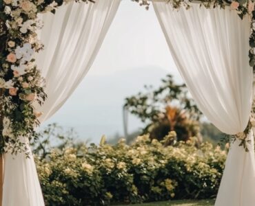 A serene outdoor wedding setup with a simple wooden arch draped in white fabric and adorned with flowers, surrounded by natural greenery under a clear sky.