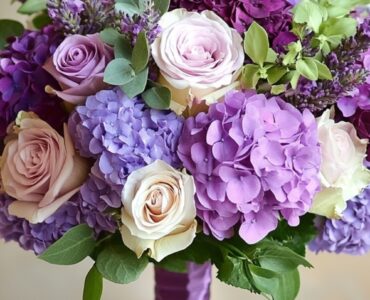 A beautifully arranged wedding bouquet featuring various purple flowers, including roses, lavender, and hydrangeas, set against a soft, romantic background.