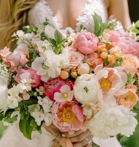A bride holding a fresh spring wedding bouquet filled with seasonal flowers in shades of pink, white, and peach, with lush greenery.